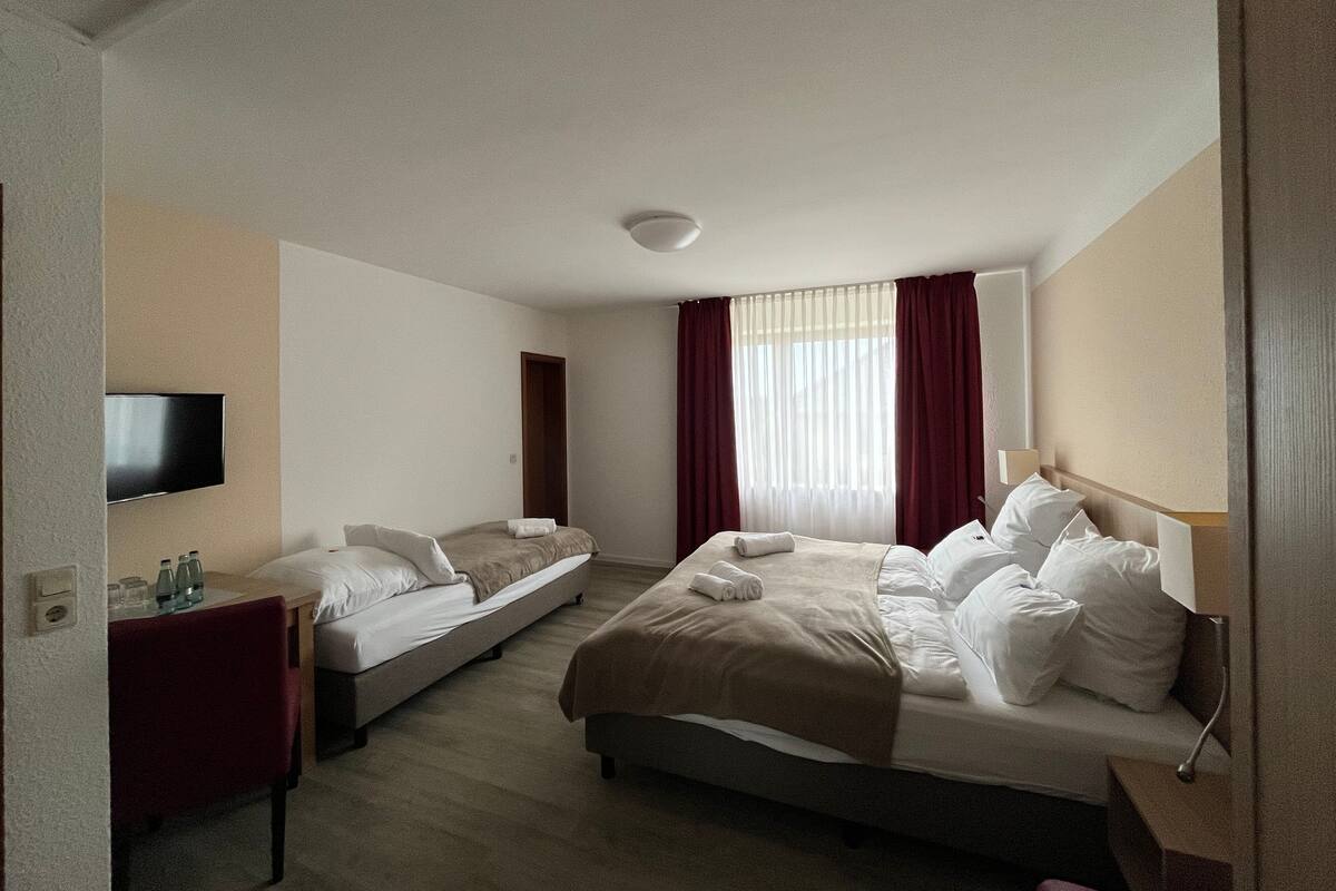 A hotel room with a large bed, a smaller bed, a desk with a chair, a TV mounted on the wall, and a window with curtains.