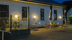 Un espace extérieur aménagé avec des tables et des chaises, une cloison en verre, et un bâtiment dont les fenêtres sont illuminées et qui porte une enseigne.