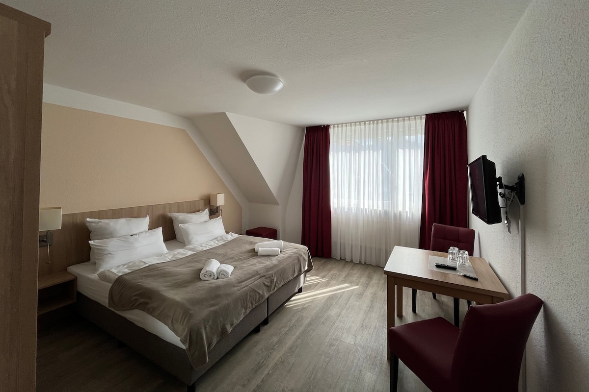 A hotel room with a bed, a desk, a chair, a TV, and a window with curtains.