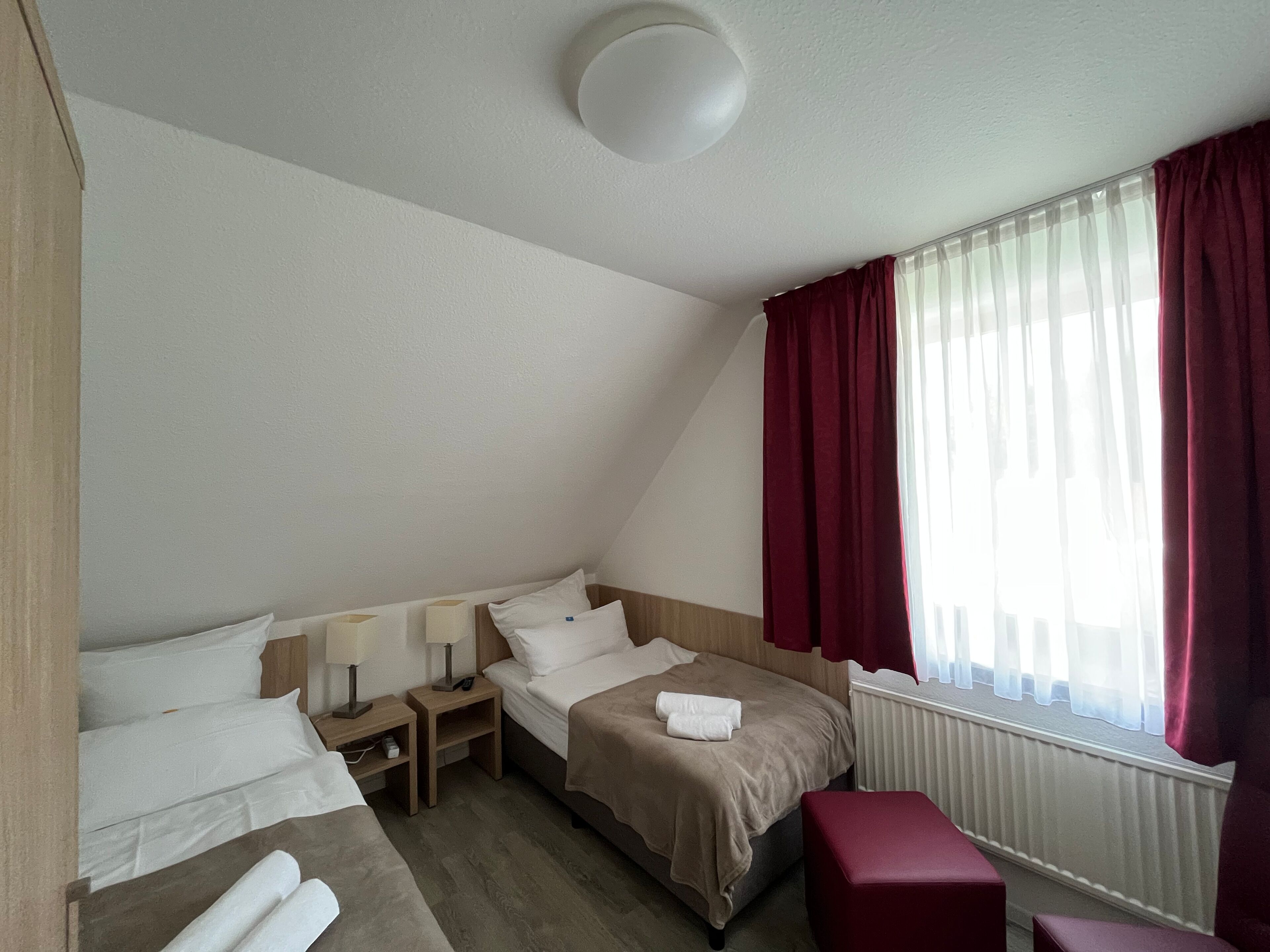 Habitación de hotel con dos camas, una mesita de noche de madera, un reposapiés rojo y una ventana con cortinas rojas.
