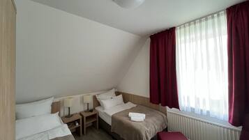 Habitación de hotel con dos camas, una mesita de noche de madera, un reposapiés rojo y una ventana con cortinas rojas.