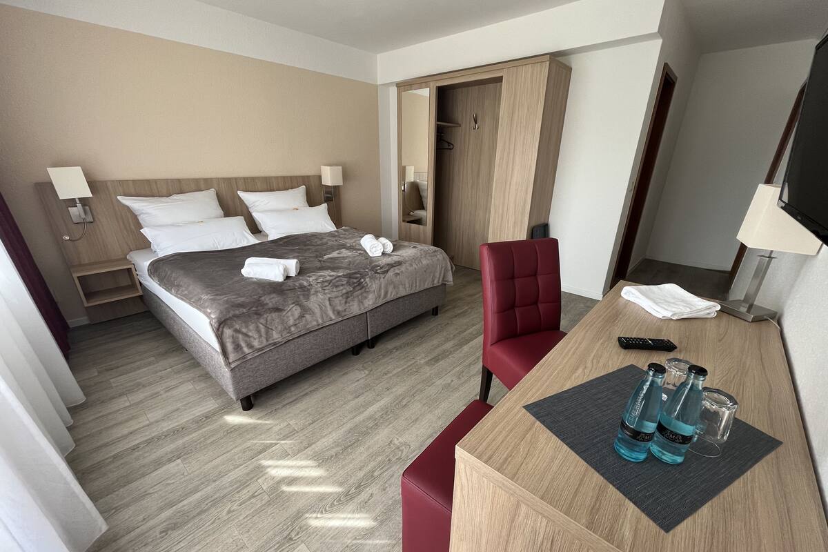 A hotel room with a bed, a desk with a chair, a TV, and a wardrobe.