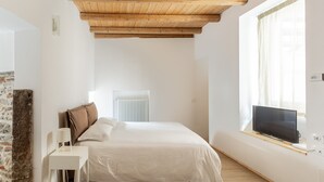 Een slaapkamer met een houten plafond, een bed met wit beddengoed, een nachtkastje met een lamp, een televisie op een plank en een raam met gordijnen.