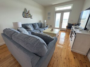 A modern living room with a grey sofa, a coffee table, and a kitchen area with a stove. - Blue Crab Condo at Rivers Edge (Steinhatchee)