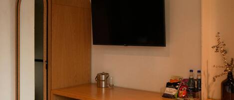 Una habitación con tocador de madera, un espejo grande, un televisor de pantalla plana y un estante con diversos objetos, incluyendo una botella, una caja y una pequeña planta.