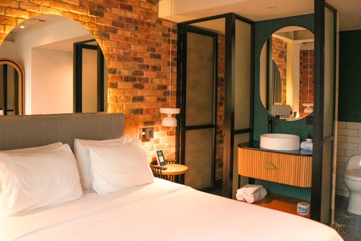A hotel room with a brick wall, a bed with white linens, a round mirror, and a bathroom with a sink and toilet.