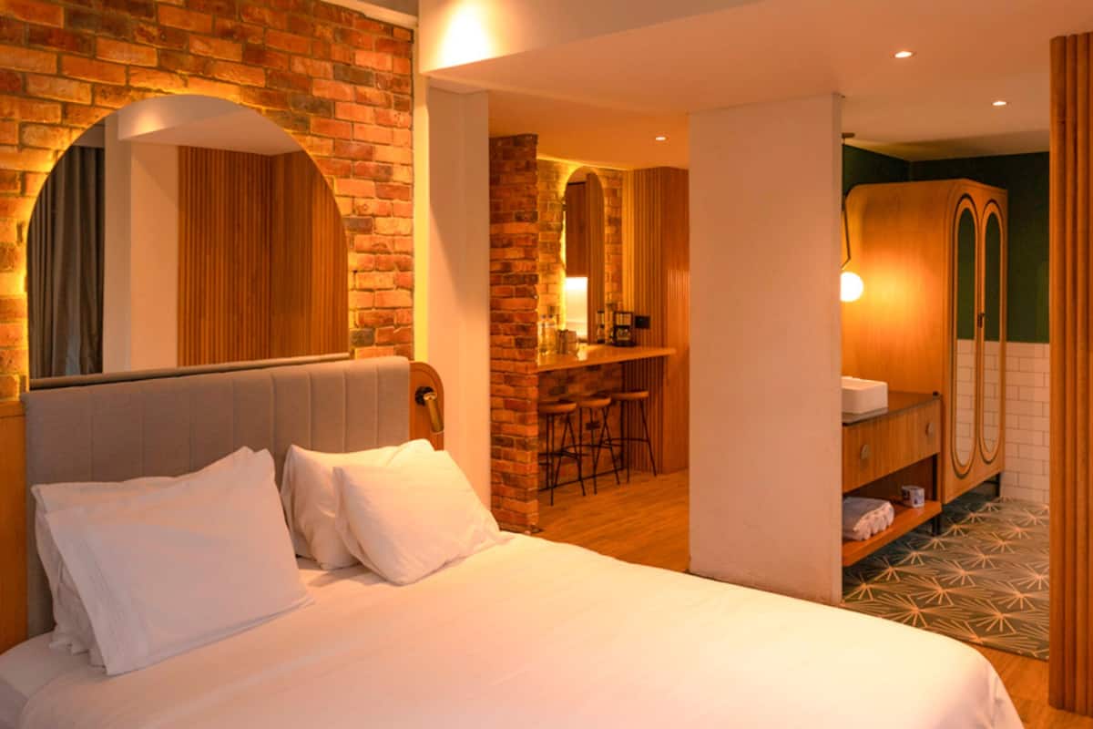 A hotel room with a large bed, a brick wall, a wooden bar, and a bathroom with a shower and sink.