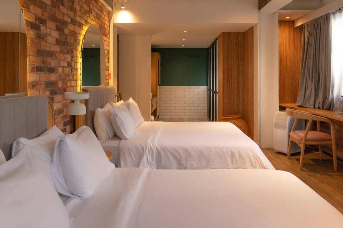 A hotel room with two beds, a desk, a chair, and a brick wall.