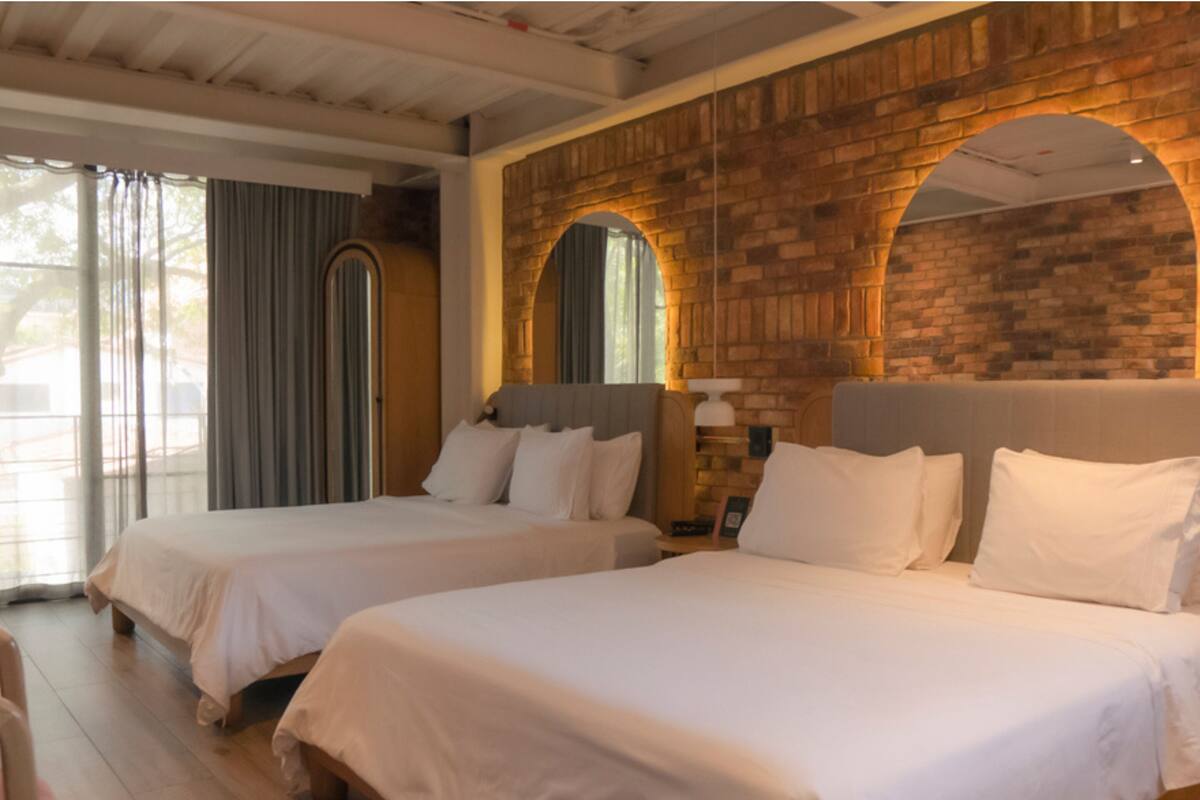A hotel room with two beds, a brick wall, and a large window with curtains.