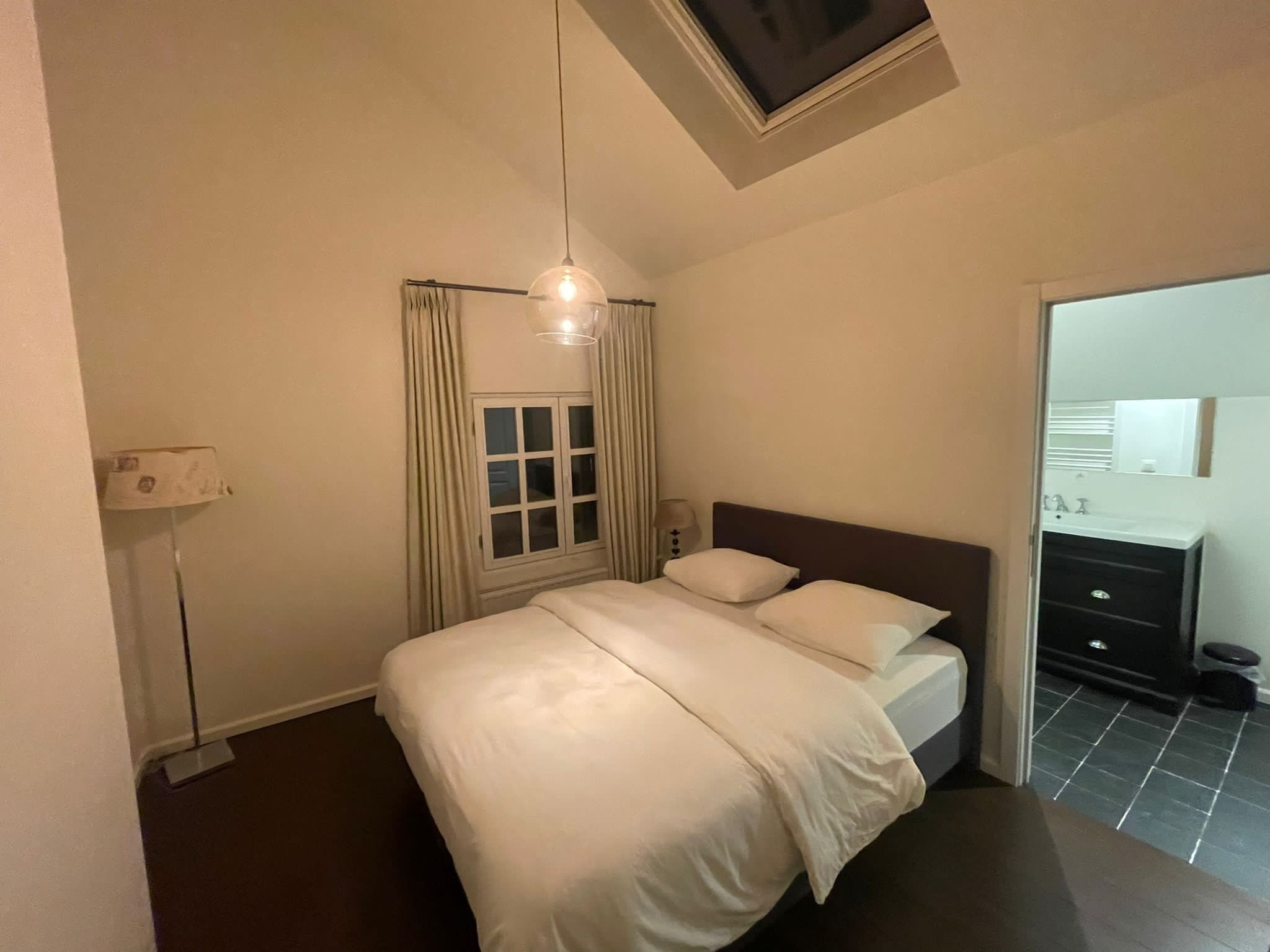 Une chambre à coucher avec un lit, une fenêtre avec des rideaux, une lampe de chevet et un luminaire au plafond.