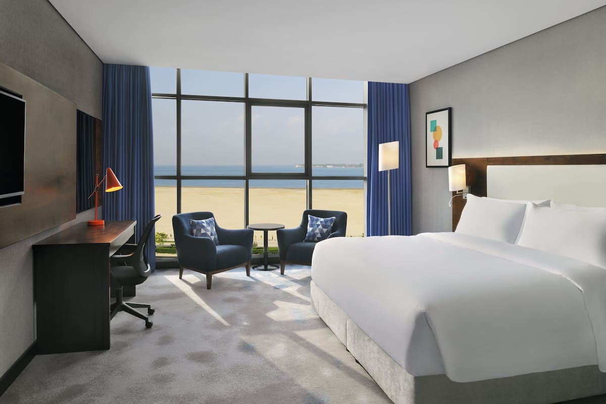 A hotel room with a large bed, a desk, two armchairs, a small table, and a view of the beach.