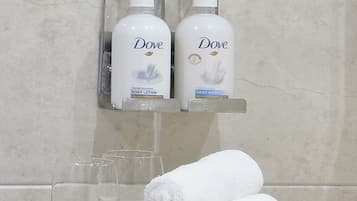 A bathroom with Dove body lotion and hand wash on a wall-mounted dispenser, a towel rack with rolled towels, and a glass of water on a tray.