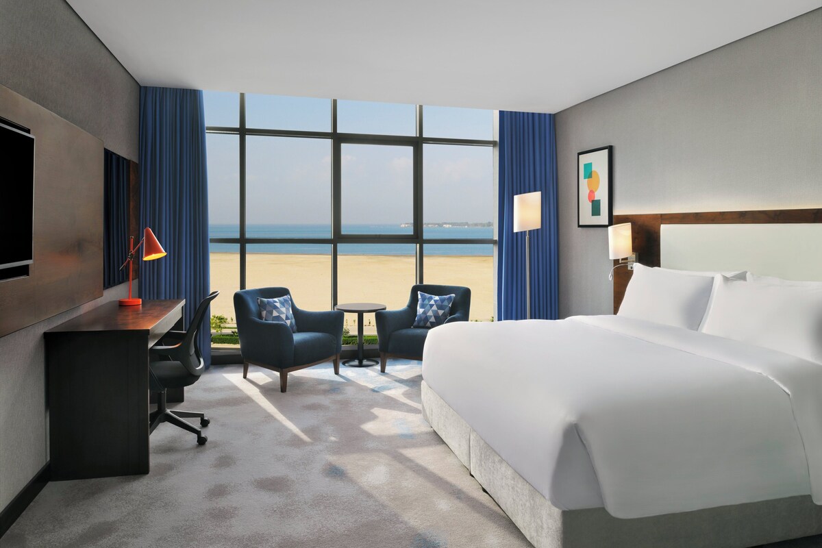 Een hotelkamer met een bed, bureau, stoelen, een televisie en uitzicht op het strand.