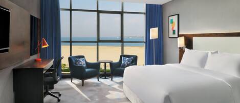 A hotel room with a bed, desk, chairs, a TV, and a view of the beach.