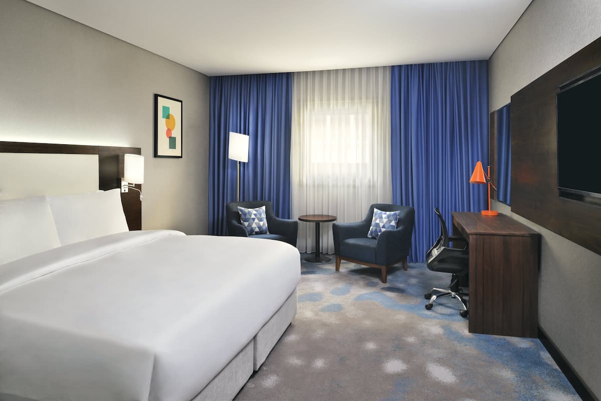 A hotel room with a large bed, two armchairs, a small round table, a desk with a lamp, and a TV.