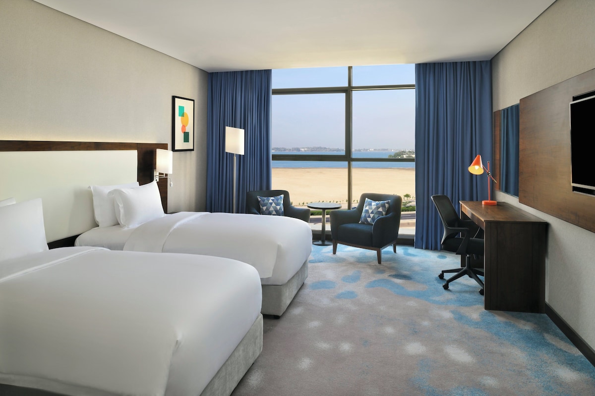 A hotel room with two beds, a desk, a chair, and a large window with a view of the beach.