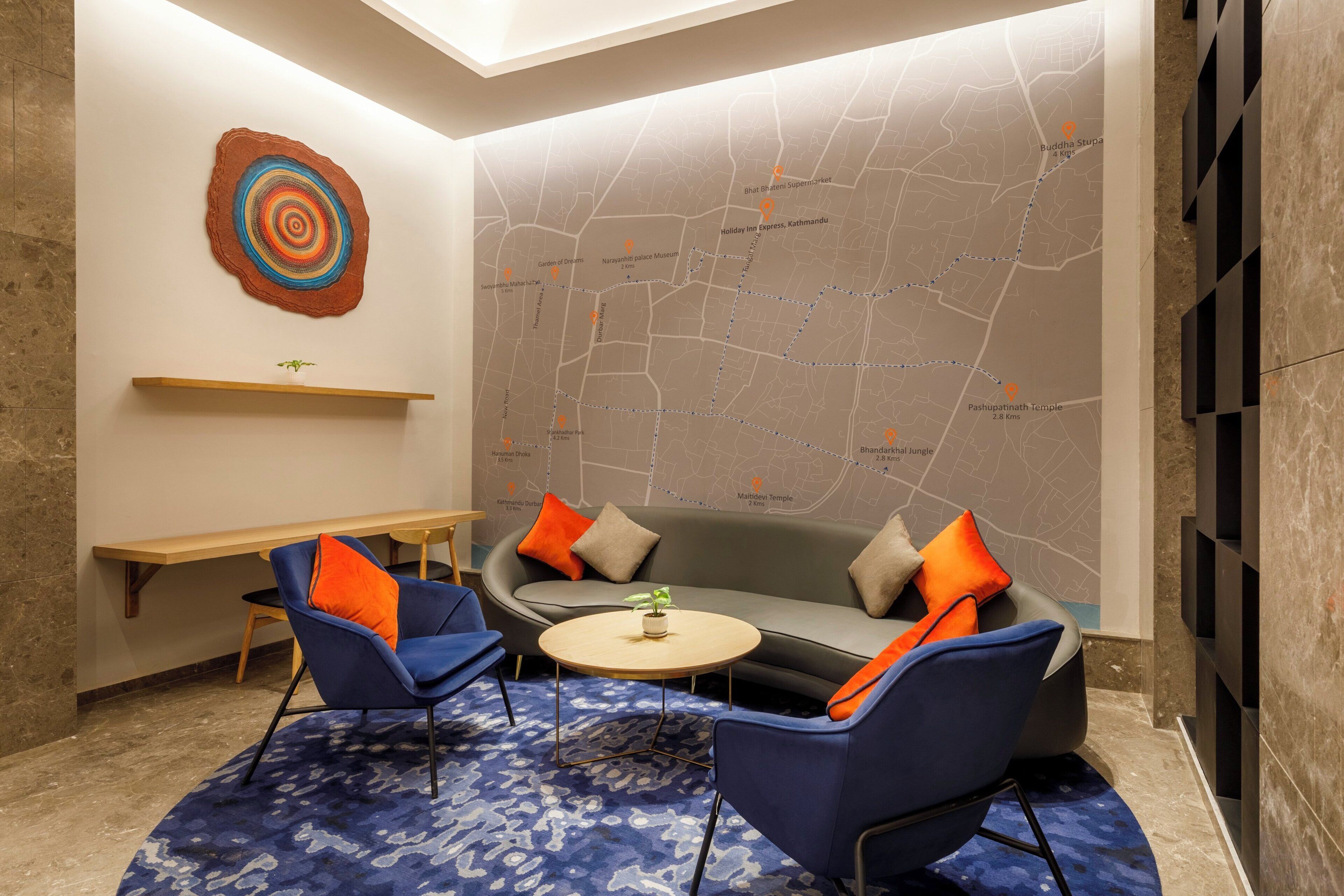 A modern lounge area with a sofa, two armchairs, a small round table, a map on the wall, and a wooden bench.