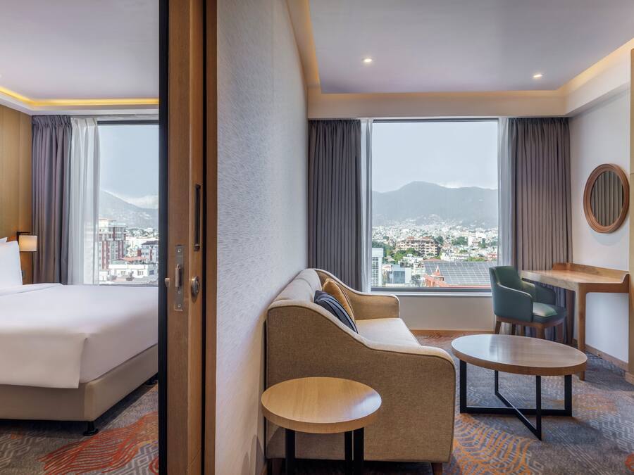 Habitación de hotel con una cama grande, un sofá, una mesita y un escritorio. Hay cortinas y vistas a un paisaje urbano con montañas al fondo.