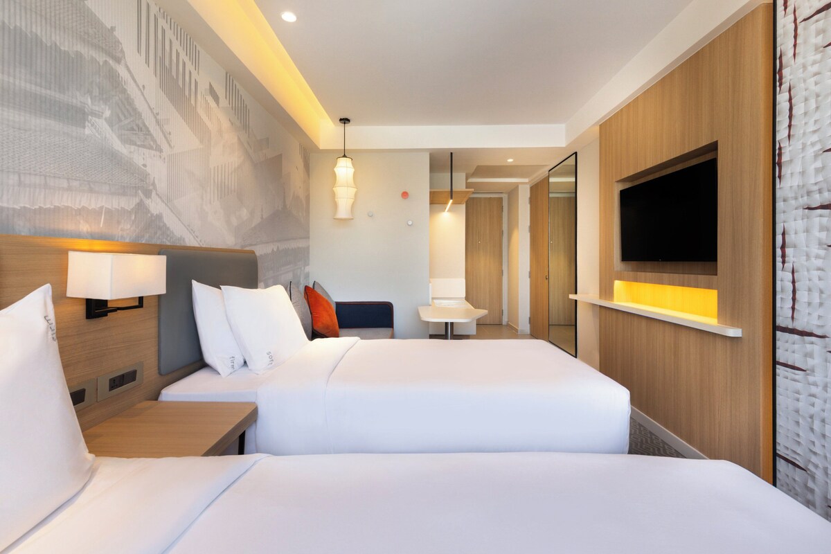 A hotel room with two beds, a TV mounted on the wall, a desk, and a large mural on the wall.