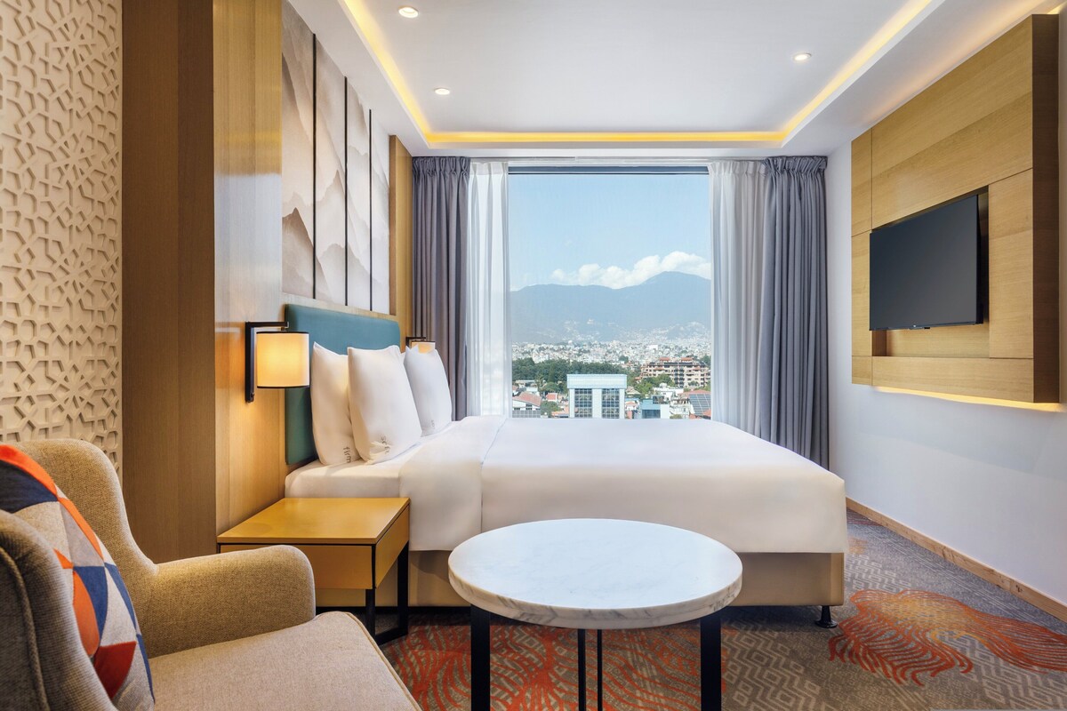 A hotel room with a large bed, a TV, a small table, and a view of a cityscape through a large window.