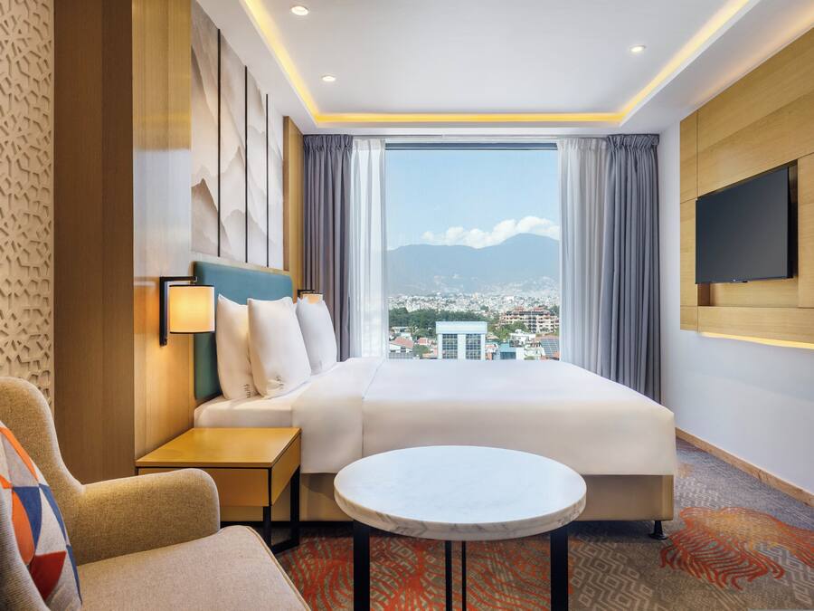 Habitació d'hotel amb un llit gran, una televisió, una taula petita i vista a una ciutat a través d'una finestra gran.