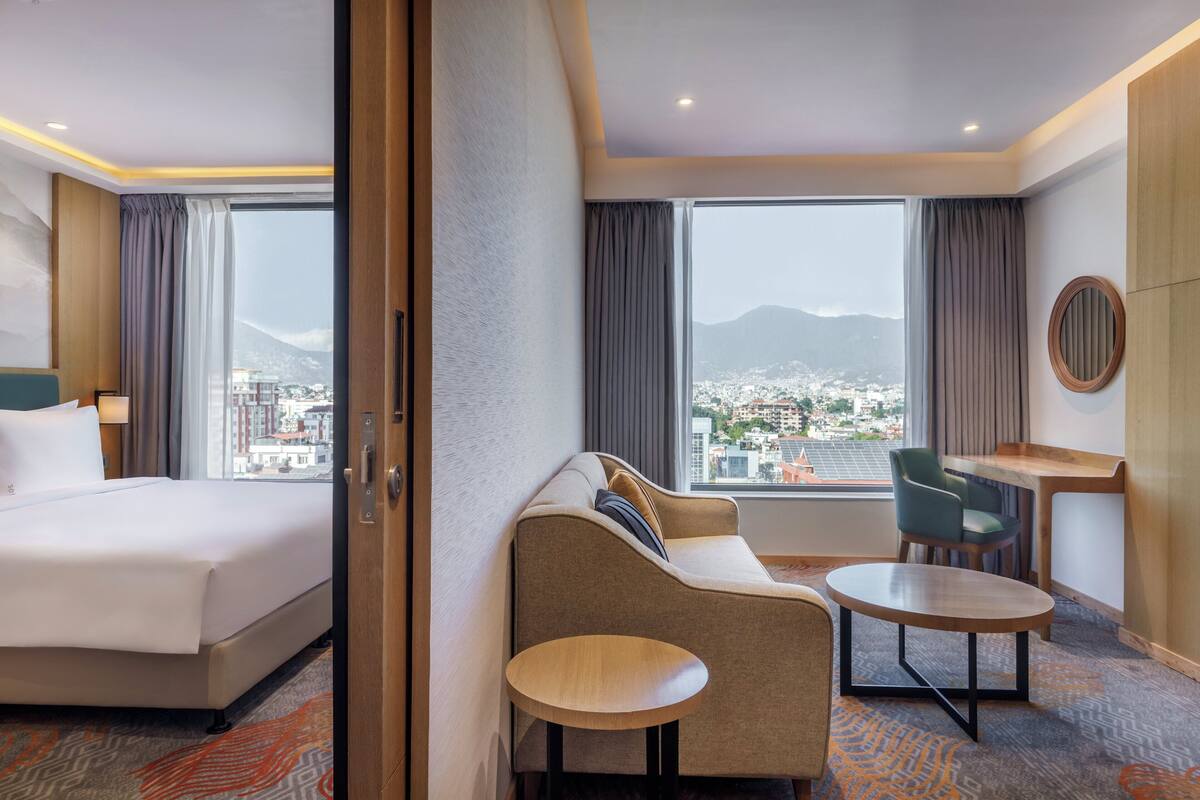 A hotel room with a large bed, a sofa, a small table, and a desk. There are curtains and a view of a cityscape with mountains in the background.
