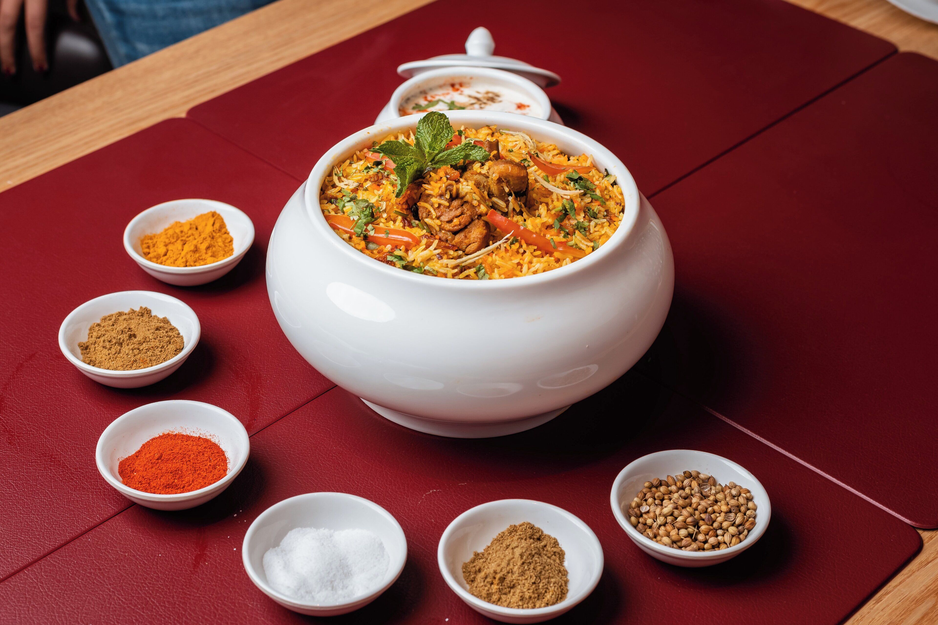 A large white pot of biryani surrounded by small bowls of spices on a red table.