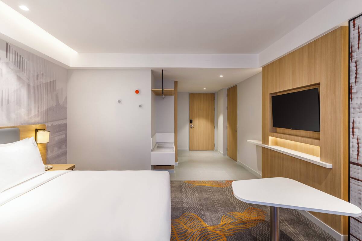 A modern hotel room with a bed, bedside table, lamp, TV, and a small desk.
