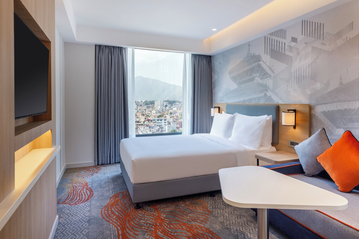 A hotel room with a bed, a desk, a TV, and a view of a cityscape.