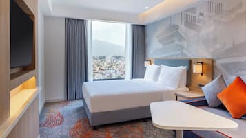 A hotel room with a bed, a desk, a TV, and a view of a cityscape.