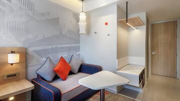 A modern hotel room with a sofa, a small table, a wall-mounted lamp, a ceiling light, and a door.