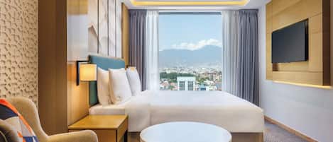 A hotel room with a bed, a desk, a chair, a TV, and a view of a cityscape.