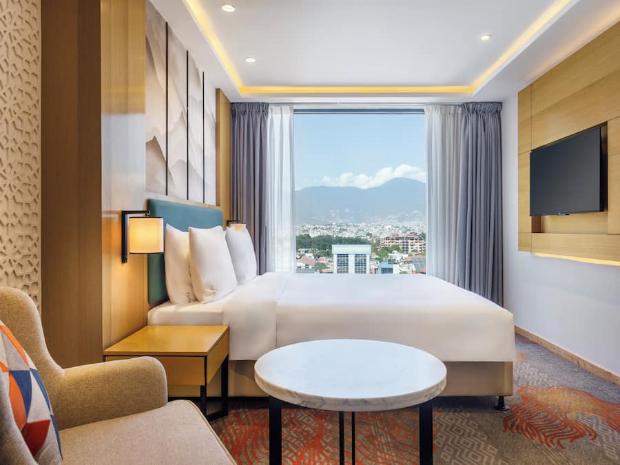 Habitación de hotel con cama, escritorio, silla, televisor y vistas a un paisaje urbano.