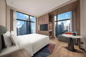 A modern hotel room with a large bed, a seating area, a flat-screen TV, and a city view.