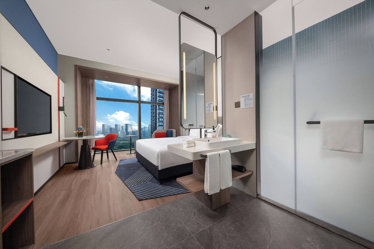 Habitación de hotel moderna con una cama grande, un escritorio, una silla, un ventanal con vistas a la ciudad, un baño con espejo y lavamanos, y una puerta.