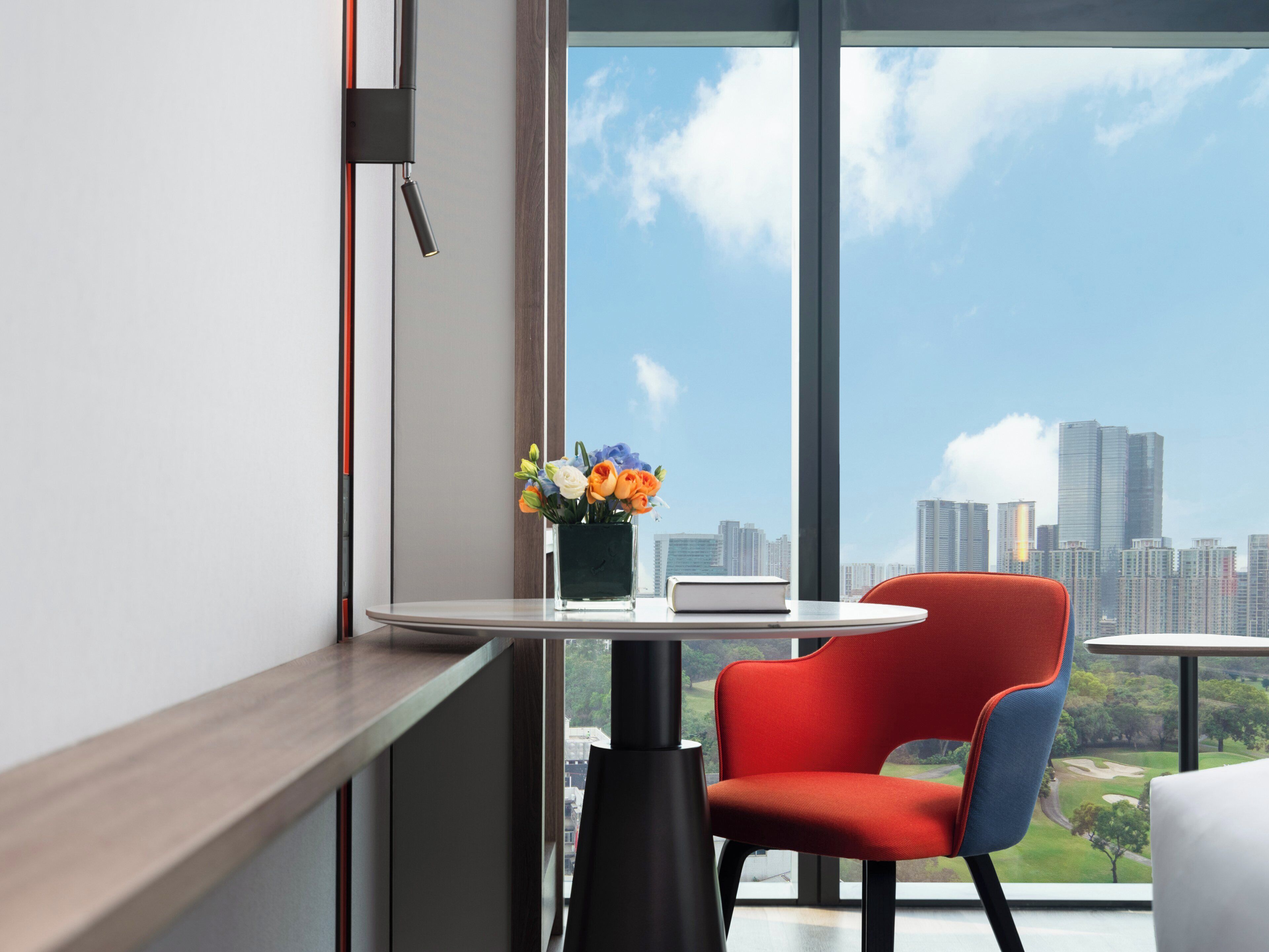 A modern hotel room with a large window offering a city skyline view, a small round table with a vase of flowers, and a red chair.