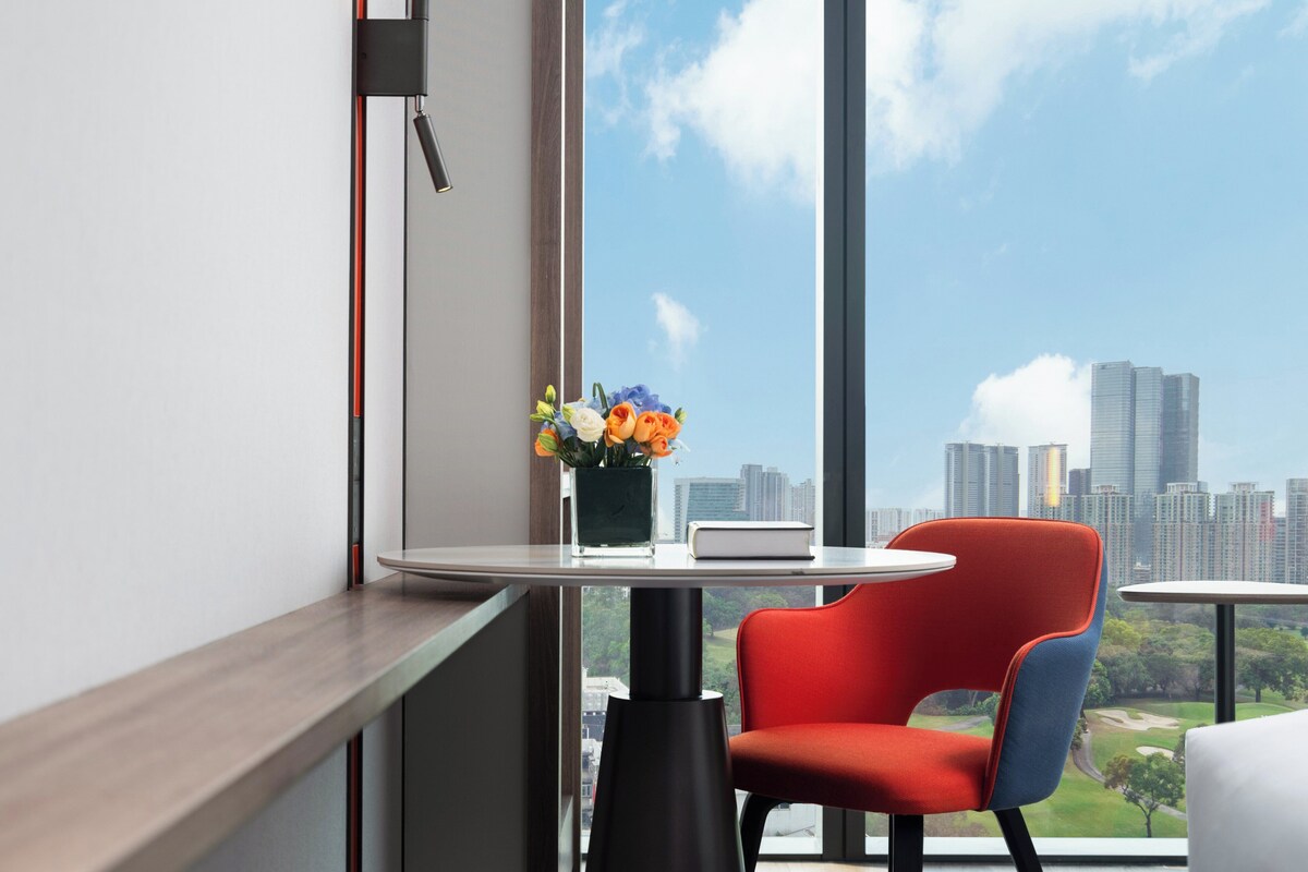 A modern hotel room with a large window offering a city skyline view, a small round table with a vase of flowers, and a red chair.