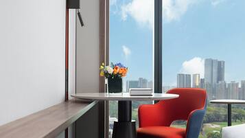 A modern hotel room with a large window offering a city skyline view, a small round table with a vase of flowers, and a red chair.