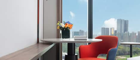 Una habitación de hotel moderna con un amplio ventanal que ofrece vistas a un horizonte urbano, una pequeña mesa redonda con un florero y una silla roja.