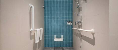 A bathroom with beige walls, a blue tiled shower area, a white grab bar, and a small white toilet.