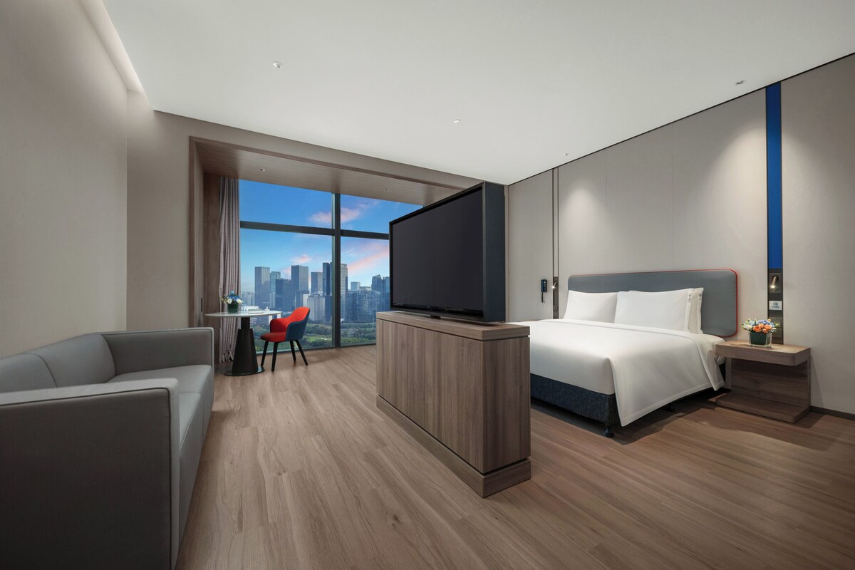 A modern hotel room with a large bed, a flat-screen TV, a seating area with a red chair, and a city view.