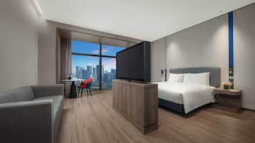 A modern hotel room with a large bed, a TV, a desk with a chair, and a view of the city skyline.