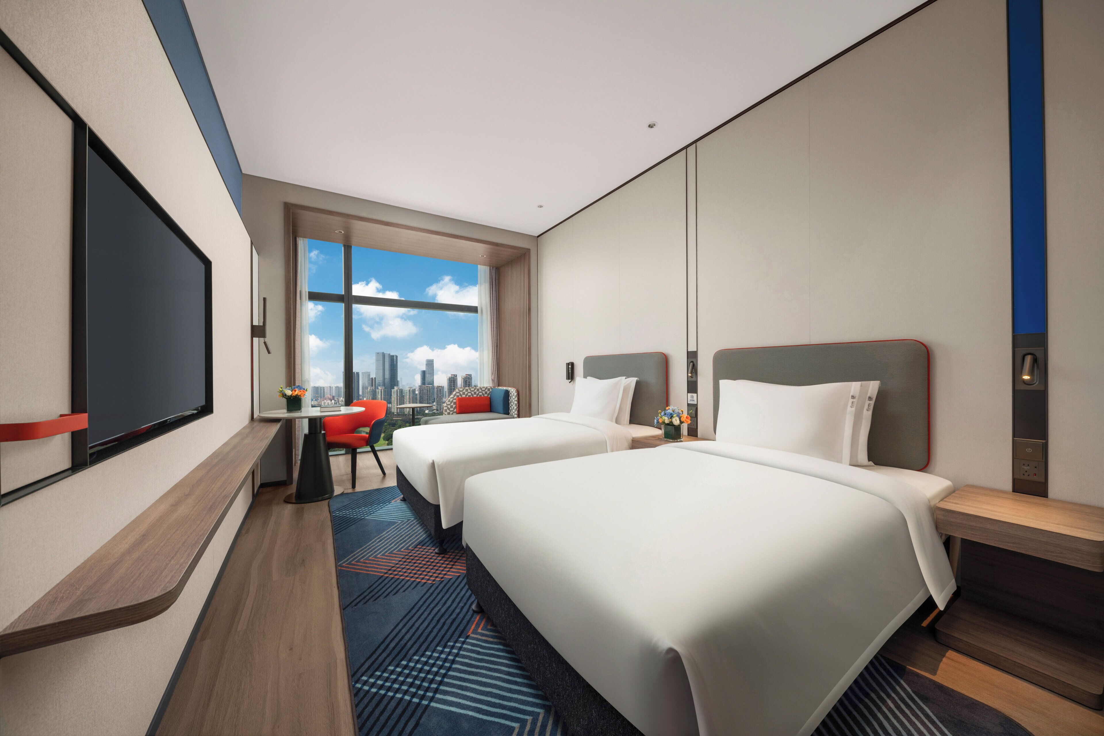 A hotel room with two beds, a TV, a city view, and a red chair.