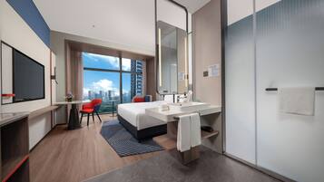 A modern hotel room with a large bed, a desk, a chair, a large window with a city view, a bathroom with a mirror and sink, and a door.
