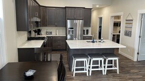 A modern kitchen with dark cabinets, a large island with bar stools, and a stainless steel refrigerator. - New Construction Home, 1 block to Legendary Surf District/ Blocks to downtown (Clear Lake)