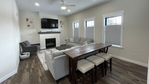 A spacious living room with a fireplace, a large flat-screen TV mounted above it, a grey sectional sofa, a wooden dining table with four barstools, and a ceiling fan. - New Construction Home, 1 block to Legendary Surf District/ Blocks to downtown (Clear Lake)