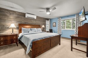 A bedroom with a wooden headboard, a bed with a blue bedspread, a nightstand with a lamp, a desk with a chair, and a ceiling fan. - Home on the Range Near Glacier Park (Columbia Falls)