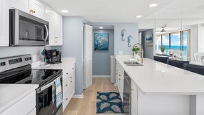 Una cocina moderna con armarios blancos, una estufa negra y una isla grande con un lavamanos. Hay un cuadro en la pared y vistas al océano a través de amplias ventanas.