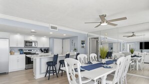 Cocina moderna con armarios blancos, una isla central y un comedor con sillas blancas y camino de mesa azul.