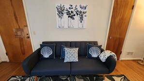 A dark blue sofa with three patterned pillows, a wall-mounted artwork of vases with leaves, and a wooden floor.